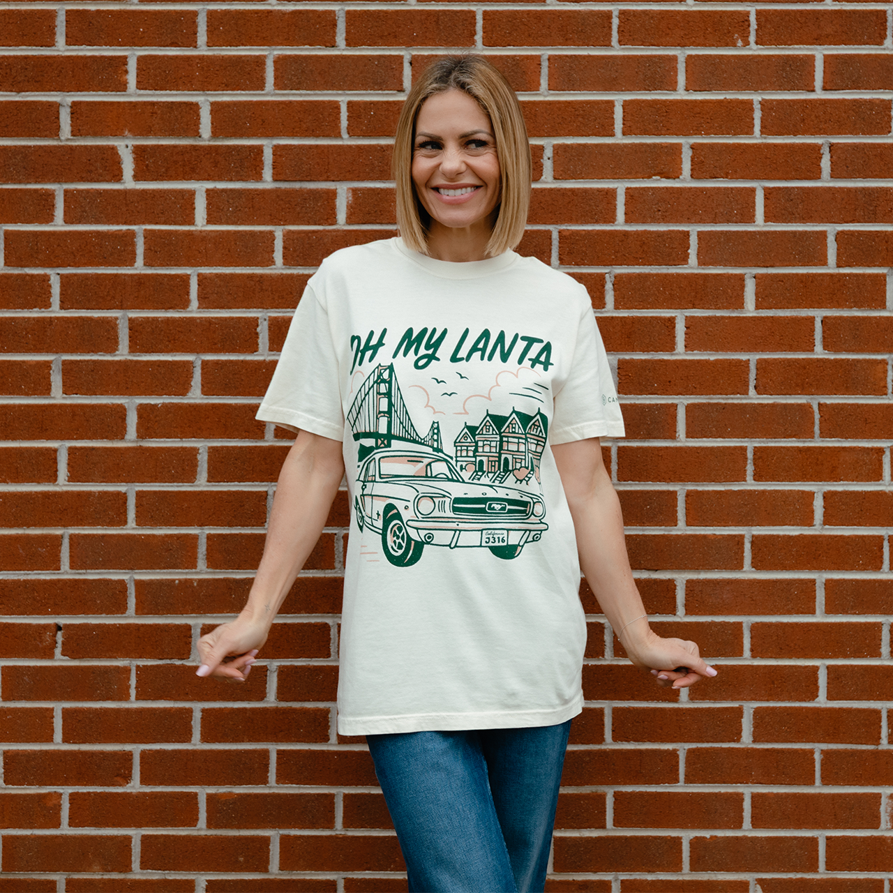 Woman wearing a white t-shirt with 'OH MY LANTA' design against a brick wall.
