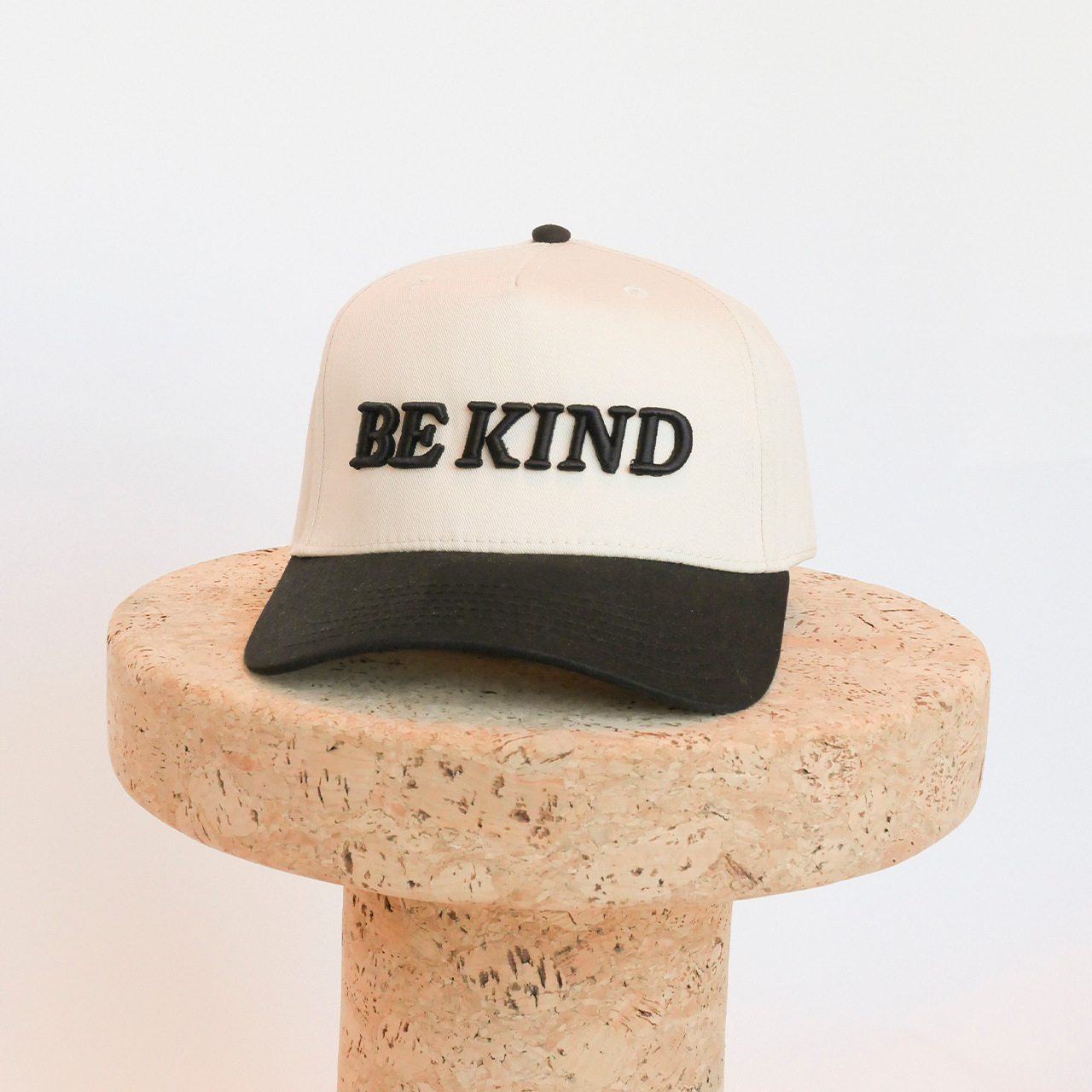 Beige cap with 'BE KIND' text on a cork block against a white background