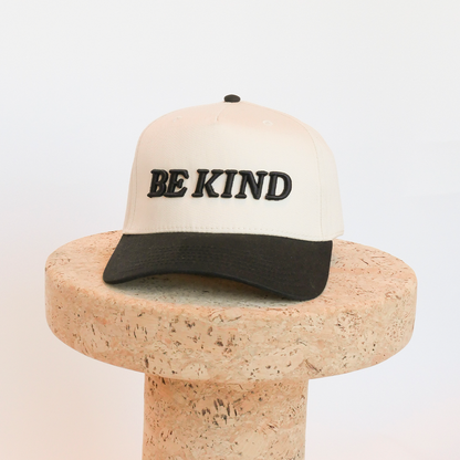 Beige cap with 'BE KIND' text on a cork block against a white background