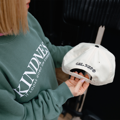 Person wearing a green sweatshirt with 'Kindness' text, holding a white cap with text.