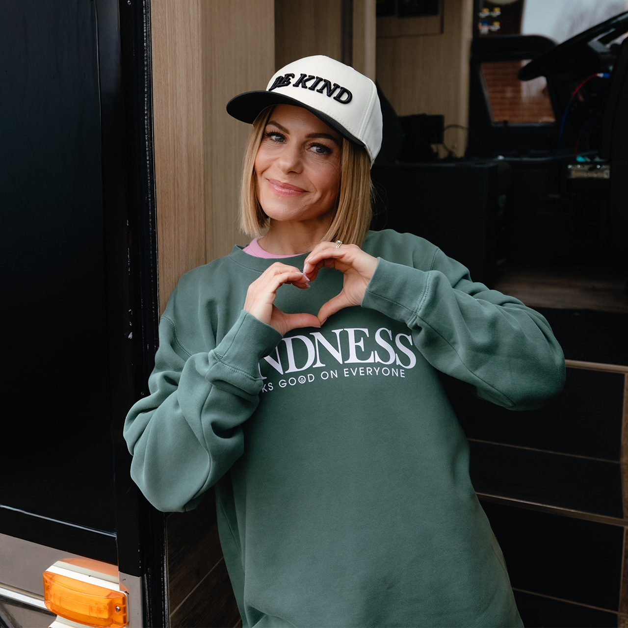 Person wearing a green sweatshirt and white cap with text, making a heart shape with their hands.