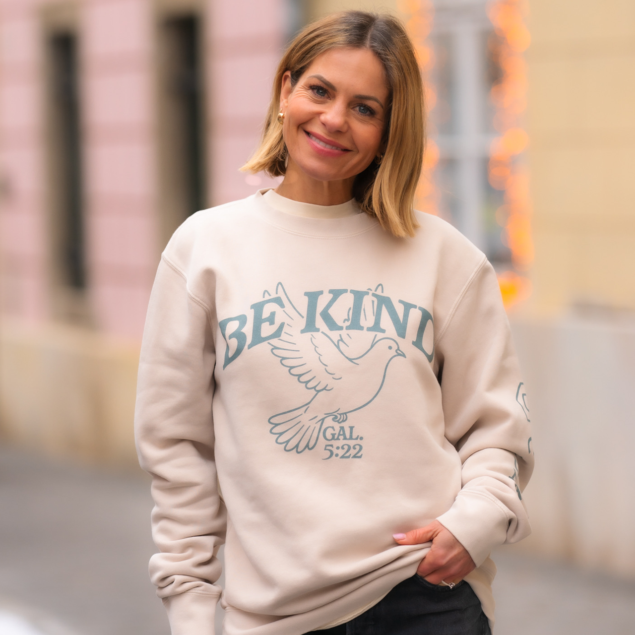 Woman wearing a beige sweatshirt with 'Be Kind' text and dove graphic, standing outdoors.